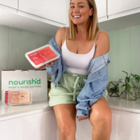 Instagram image of a young woman sitting at a small dining table eating her Nourish'd fresh meal from a bow, with a crystal glass of kombucha beside her.